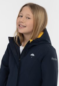 Marinblå jacka med gul foder, med dragkedja och kardborrestängning. Inkluderar en silvrig paraplylogotyp och varumärkesnamn på ärmen.