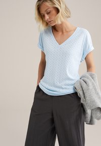 Gaiši zilā V-veida blūze ar teksturētu zvīņveida rakstu, īsām rullētām piedurknēm. Apvienota ar tumšiem, platām kājām biksēm un pelēku adījumu.