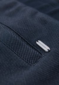 Tissu texturé bleu foncé avec une petite étiquette métallique indiquant "QUINTESSENCE" attachée près d'une couture ou du bord d'une poche.