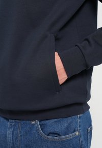 Marineblauwe sweatshirt met zijzak en geribde manchetten. De stof lijkt glad en zacht. Gekleed met blauwe spijkerbroek.