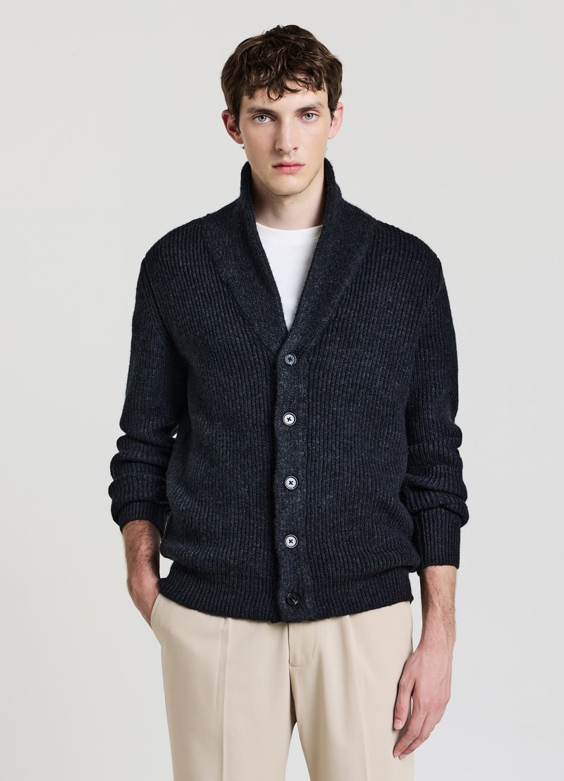 Cardigan grigio scuro a coste con collo sciallato, maniche lunghe e cinque bottoni. Indossato sopra una camicia bianca, abbinato a pantaloni beige chiaro.
