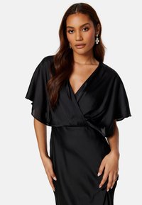 Zwarte satijnen jurk met een diepe V-hals en korte fluttermouwen. Zachte textuur en getailleerde silhouet. Vrouw poseert met handen op de heupen.