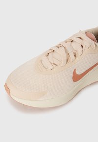 Helle beigefarbene Sportschuh mit einem strukturierten Mesh-Obermaterial, flachen Schnürsenkeln und einem dezenten Koralle-Akzent am Swoosh und der Zehenkappe. Gummisohle.