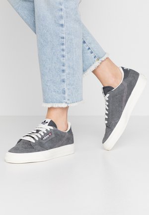 Personne portant des baskets en suède Adidas grises avec des lacets blancs et un jean bleu clair à ourlet effiloché sur un fond clair uni.