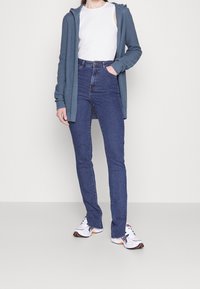 Lichtblauwe gebreide cardigan met capuchon, witte tanktop, high-waist donkerblauwe jeans en witte sneakers met oranje accenten.