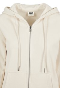 Sweat à capuche zippé couleur crème avec cordons, poches avant et étiquette Urban Classics Organic à l'intérieur du col.