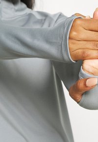 Jasnoszara koszulka z długim rękawem i zamkiem przy kołnierzu, wykonana z gładkiego, elastycznego materiału z otworami na kciuki dla dodatkowego komfortu i osłony.