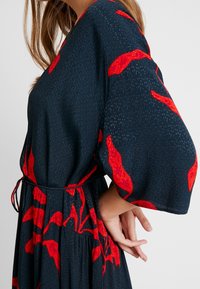 Šaty s květinovým vzorem na tmavě modrém pozadí, s živými červenými akcenty, ¾ rukávy a zavazováním v pase. Texturovaná látka.