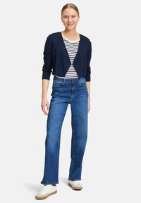 Cardigan corto blu sopra una camicia a righe bianche e blu, abbinato a jeans dritti blu e sneaker bianche con suola beige.