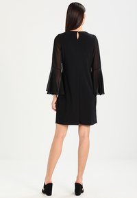 Robe noire avec des manches évasées transparentes et un détail de dos en trou de serrure. Longueur courte avec une texture lisse et un design ajusté.