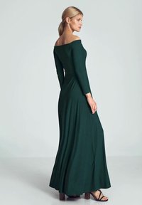 Lang groen off-shoulder jurk met lange mouwen, aansluitend lijfje en vloeiende rok, voorzien van een gestructureerde stof en zwarte hoge hakken.