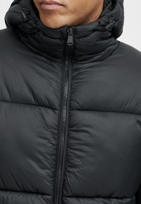 Chaqueta acolchada negra con un cuello alto, que presenta secciones acolchadas visibles, un cierre central y cordones ajustables en la capucha.
