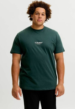 Junger Mann mit lockigem Haar und Sommersprossen, der ein dunkelgrünes JJ Studios T-Shirt trägt und vor einem schlichten hellen Hintergrund steht.