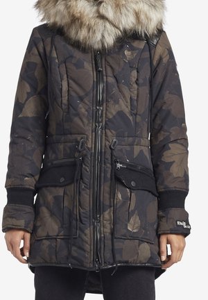 Tmavý zimní kabát s květinovým vzorem, s kapucí lemovanou kožešinou, zapínáním na zip a knoflíky a velkými předními kapsami, na osobě stojící nehybně.