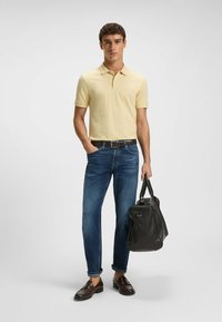 Gul pikétröja, blå denimjeans, brun bältet, mörka loafers, håller en svart läderväska. Enkel design och avslappnad look.