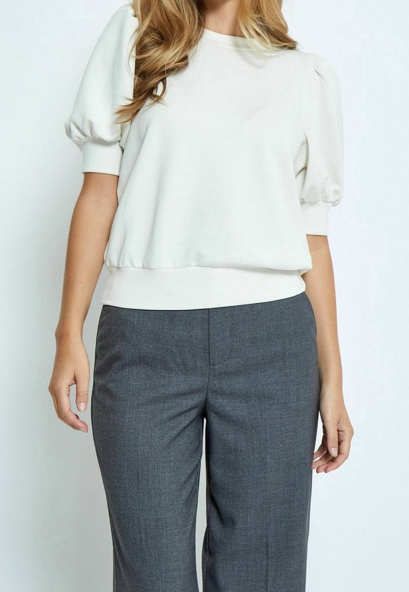 Femme portant un chemisier blanc à manches bouffantes et un pantalon gris sur mesure, debout devant un fond clair uni.