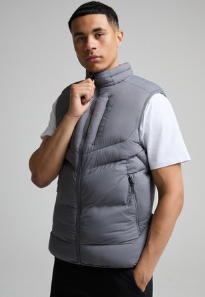 Ung mand med kort hår iført en grå polstret vest over en hvid kortærmet skjorte, der lynlåser vesten med den ene hånd.