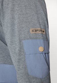 Gray, textured fabric shirt featuring a blue pocket with a rounded, tan button and a leather logo patch labeled "ICEPEAK."