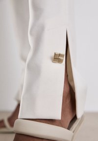 Pantalon blanc ajusté avec un ourlet fendu, arborant un accent en métal doré en forme de carré. Le tissu a une texture lisse et une finition soignée.