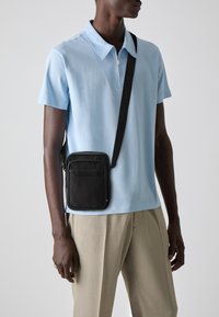 Geantă de umăr neagră, cu textură netedă și buzunar cu fermoar, purtată peste un tricou polo de un albastru deschis. Geanta are o curea cu un logo.