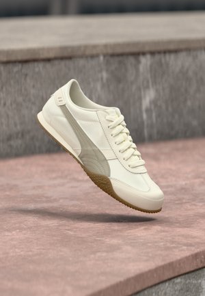 Off-white sneaker med lysegrå stribe og gummisål, vist i luften over stenetrin udendørs.