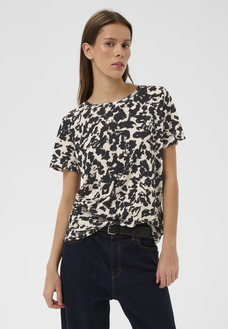 Junge Frau mit langen braunen Haaren, die ein kurzärmeliges Shirt mit abstraktem Schwarz-Weiß-Muster und dunkle Jeans mit schwarzem Gürtel trägt.