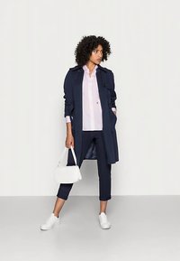 Marineblauwe trenchcoat met een ceintuur in de taille, gedragen over een lichtroze shirt, donkergroene broek, witte sneakers en een klein wit handtasje. Soepele texturen.