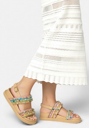Beżowe sandały na platformie z wielokolorowymi, cekinowymi paskami, noszone z białą, teksturowaną spódnicą midi.