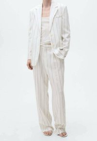 Costume blanc à fines rayures avec une veste croisée à un seul bouton et un pantalon à jambes larges. Le tissu semble léger, avec une texture subtile ; porté avec un haut ajusté.