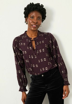 Purple blouse with gold floral patterns, ruffled neckline, and ties; long sleeves with elastic cuffs. Paired with black pants.