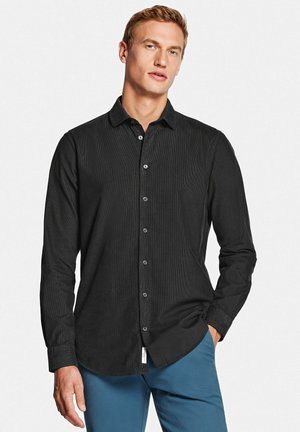 Jonge man met kort blond haar, gekleed in een zwart overhemd met knopen en een blauwe broek, staand met één hand in de zak tegen een witte achtergrond.