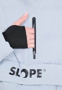 Vaaleansininen eristetty takki, jossa on musta hihansuu peukalonreiällä, sivuzippitasku ja suuri musta "SLOPE" -logo alaosassa.