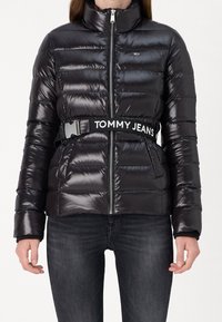 Svart vadderad jacka med hög krage, horisontell sömnad, dragkedja och ett märkesbälte. Har elastiska muddar och sidofickor.