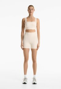 Beige sportieve crop top met vierkante halslijn en gedetailleerde banden, gecombineerd met hoge taille bijpassende shorts, gemaakt van nauwsluitend, rekbaar materiaal.