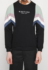 Sudadera negra con rayas en los hombros de varios colores en verde menta, rosa, blanco y azul marino. Presenta el logo "KAOTIKO" en el pecho.