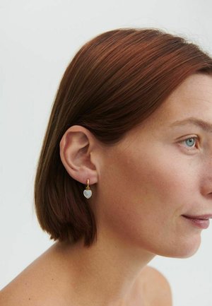 Sieviete ar īsiem sarkaniem matiem, valkājot mazus zelta stīpas auskarus ar baltām sirds formas kuloniem, skatās pa labi pret vienkāršu fonu.