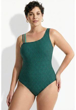 Kvinde med kort mørkt hår iført grøn og sort ternet badedragt med én skulder og dobbelte tynde stropper på hvid baggrund.