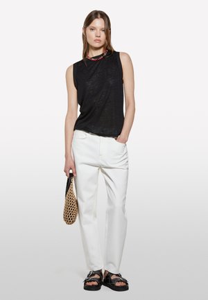 Top senza maniche nero, pantaloni bianchi, sandali neri, che tiene una borsa intrecciata. Design semplice, vestibilità rilassata e dettagli in tessuto testurizzato.