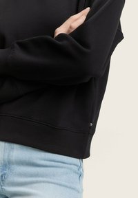 Felpa nera con colletto rotondo a coste, maniche lunghe e un orlo ben definito con un piccolo etichetta del logo, abbinata a jeans azzurri chiari.