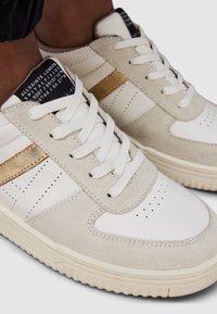 Białe i beżowe sneakersy wykonane z zamszu i skóry, z złotym akcentem na pasku, białymi sznurowadłami oraz perforacjami na przedniej części.