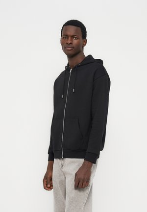 Mann iført en svart hettegenser med glidelås og snorer, og lysegrå jeans, stående mot en ensfarget hvit bakgrunn.