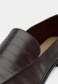 Mocassin en cuir bordeaux avec un motif texturé croco, bout arrondi et coutures contrastantes. Semelle en caoutchouc noire.