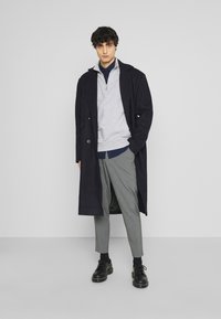 Lungo cappotto blu navy sopra un maglione grigio, stratificato con una camicia blu navy, abbinato a pantaloni a quadri grigi e scarpe nere. Texture lisce in tutto.