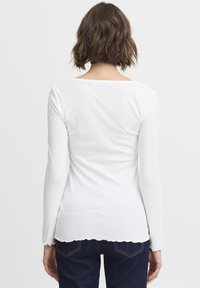 Langärmliges weißes Rippoberteil mit einem runden Rückenausschnitt, gewelltem Saum und figurnaher Passform, kombiniert mit dunklen Jeans.