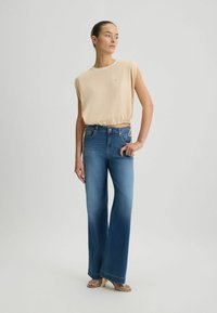 Top beige a maniche corte con orlo regolabile, abbinato a jeans in denim a vita alta con gamba larga, che sfumano dal blu scuro al blu chiaro.