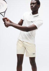 Λευκό πόλο με μαύρο περίγραμμα και λογότυπο, συνδυασμένο με ανοιχτό μπεζ σορτς που φέρει επίσης λογότυπο. Το μοντέλο κρατά ρακέτα τένις.