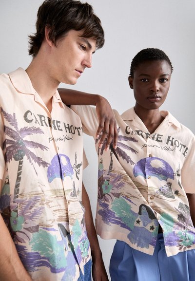 Short-sleeve, button-up shirts in light peach with tropical print featuring palm trees, water, and abstract figures. Text reads "CARNE HOTE pura Vida."
