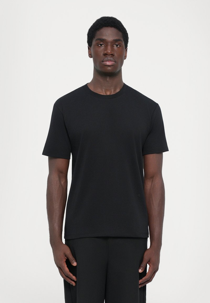 Schwarzes kurzärmeliges T-Shirt aus glattem Stoff, mit einem klassischen Rundhalsausschnitt und geradem Schnitt, getragen mit schwarzen Hosen. Minimalistisches Design.