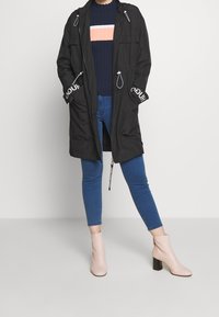 Parka negra con capucha ajustable, acentos de logo blanco en las mangas, combinada con un suéter de canalé azul marino y jeans ajustados azules, usando botas de tacón rosas claras.