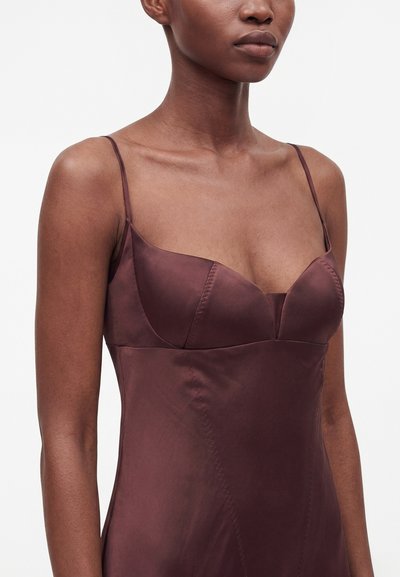 Femme portant une robe en satin marron foncé sans manches avec des fines bretelles et un détail ajusté au niveau de la poitrine, montrée des épaules jusqu'au milieu du torse.